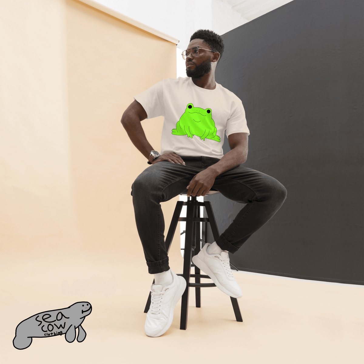 Person sitting on a stool wearing a white t-shirt with a green frog graphic, black pants, and white sneakers.