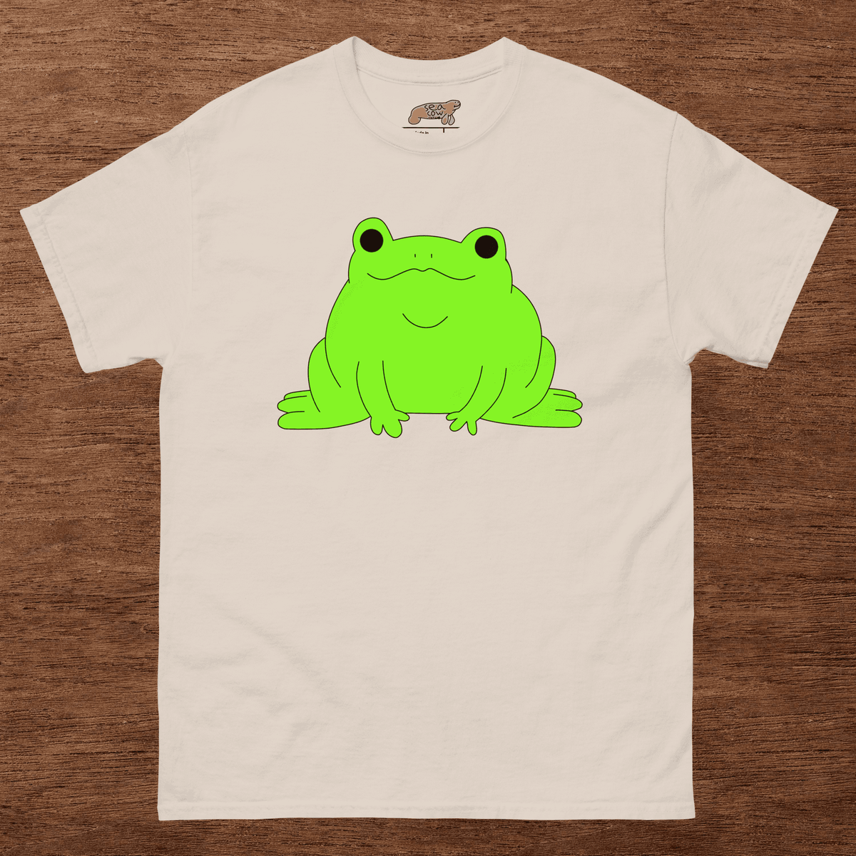 Beige t-shirt with a green frog graphic on a wooden surface