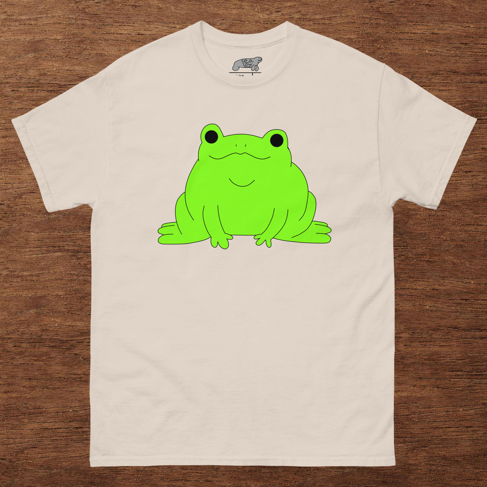 Beige t-shirt with a green frog graphic on a wooden surface